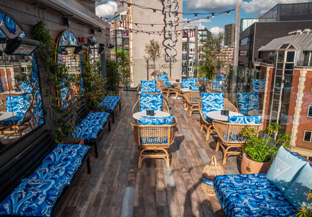 Virgin Hotels outdoor patio with wicker seating, vibrant blue cushions, plants, and city views, perfect for relaxed lounging.