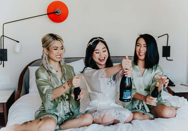 Bride and bridesmaids sitting on a signature Virgin Hotels bed popping champagne