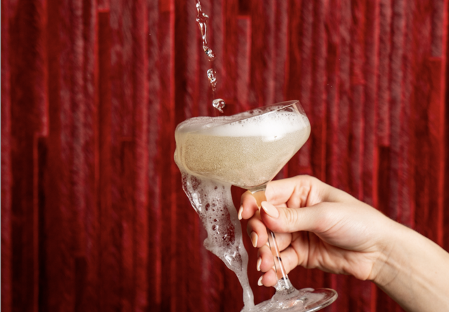 Champagne being poured over a full glass with a red curtain backdrop.