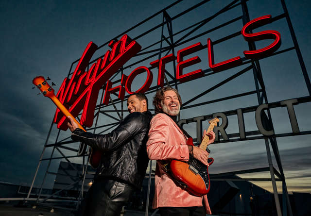 Two guistarists posing back to back in front of a sign of Virgin Hotels Dallas Design District