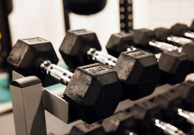 A gym rack with black hex dumbbells neatly arranged on a silver stand, reflecting light in a well-equipped Virgin Hotels fitness center.