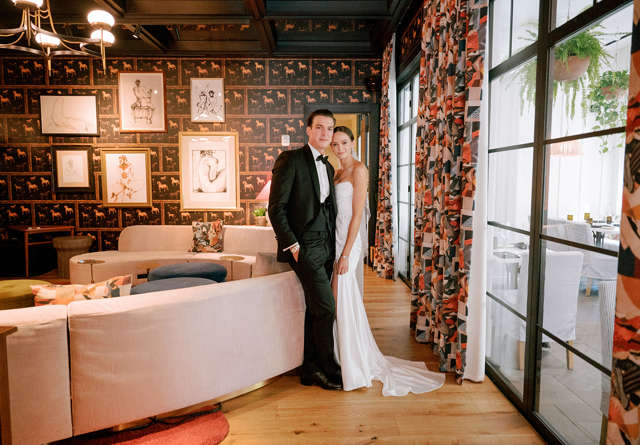 A bride and groom pose together in a stylish lounge, featuring elegant decor and colorful patterned curtains.